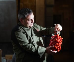 Chef Nunzio Saviano tiene in mano un grappolo di pomodorini del piennolo del Vesuvio in un ambiente rustico, simbolo della tradizione campana.
